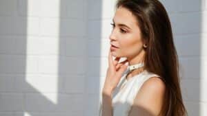 Woman with clear glowing skin touching her face, sunlight on white brick wall background.